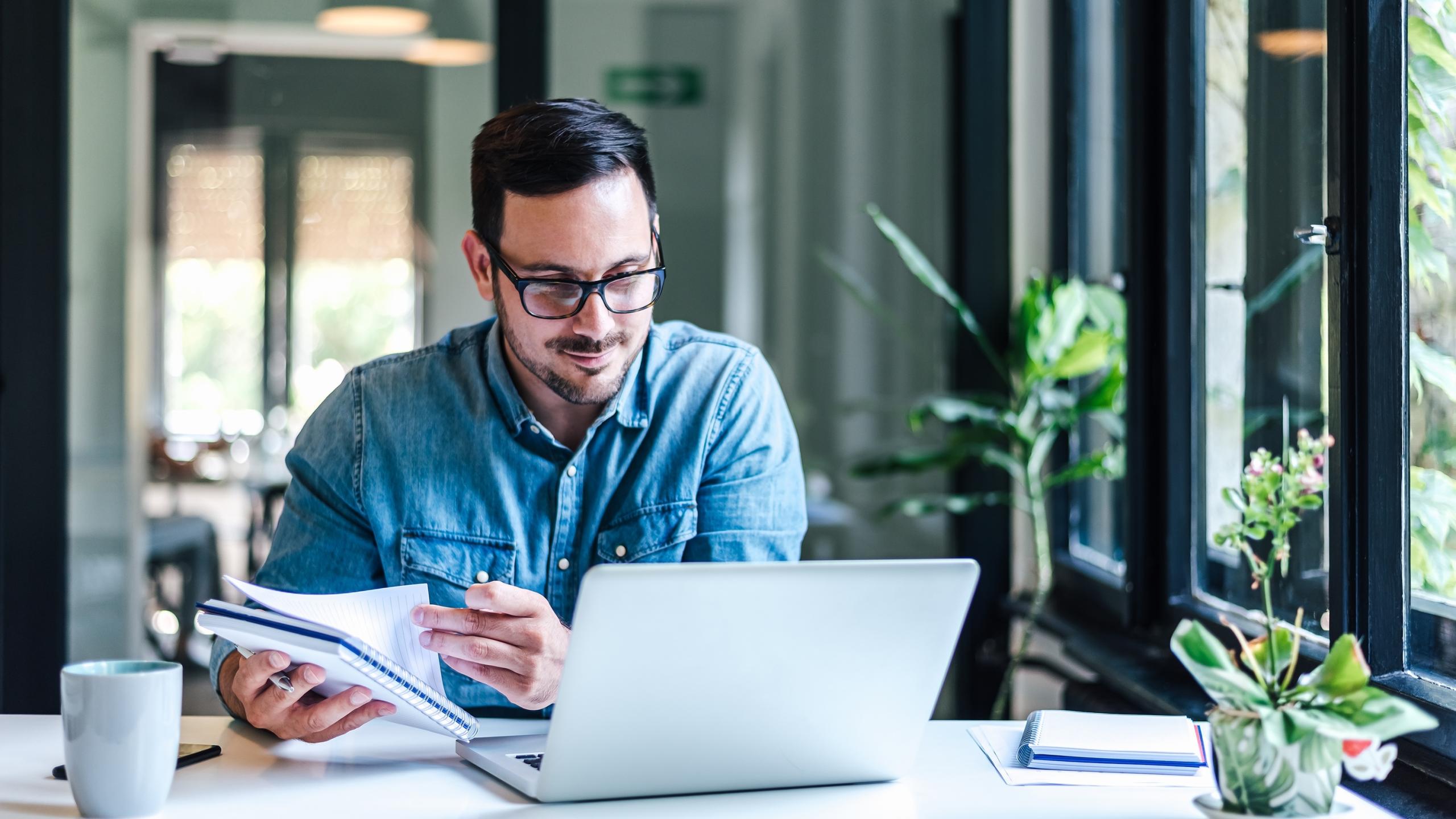 Blije dertiger in jeanshemd aan bureau met laptop.