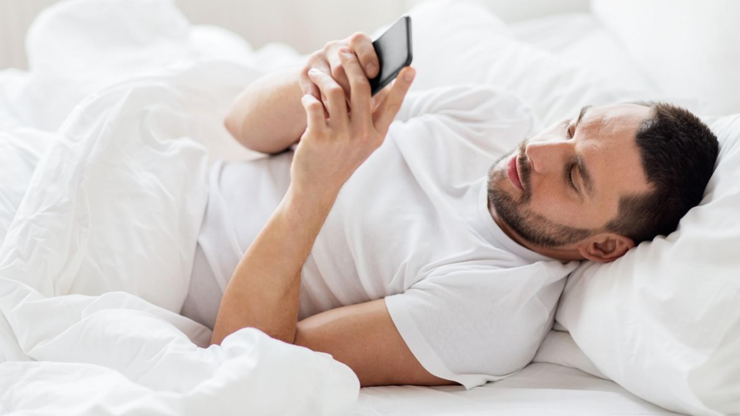 Man in bed kijkt naar smartphone.